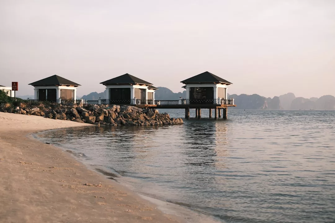 Three small pavilions on stilts stand over calm water near a sandy beach, offering a taste of luxury lifestyle against Majorca’s stunning rocky coastline and distant mountains.