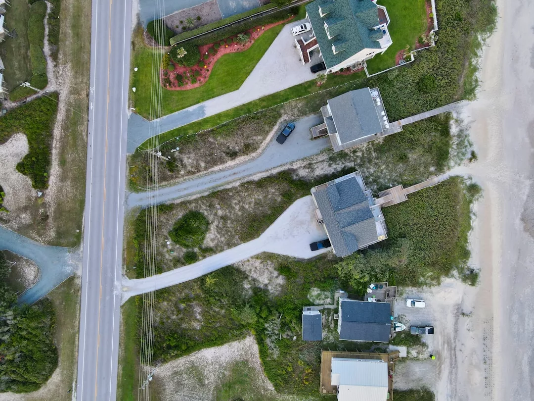 Aerial view of a luxury coastal neighborhood in Majorca, featuring elegant houses, driveways, a main road, lush grassy areas, and a sandy beach that perfectly captures the island lifestyle.