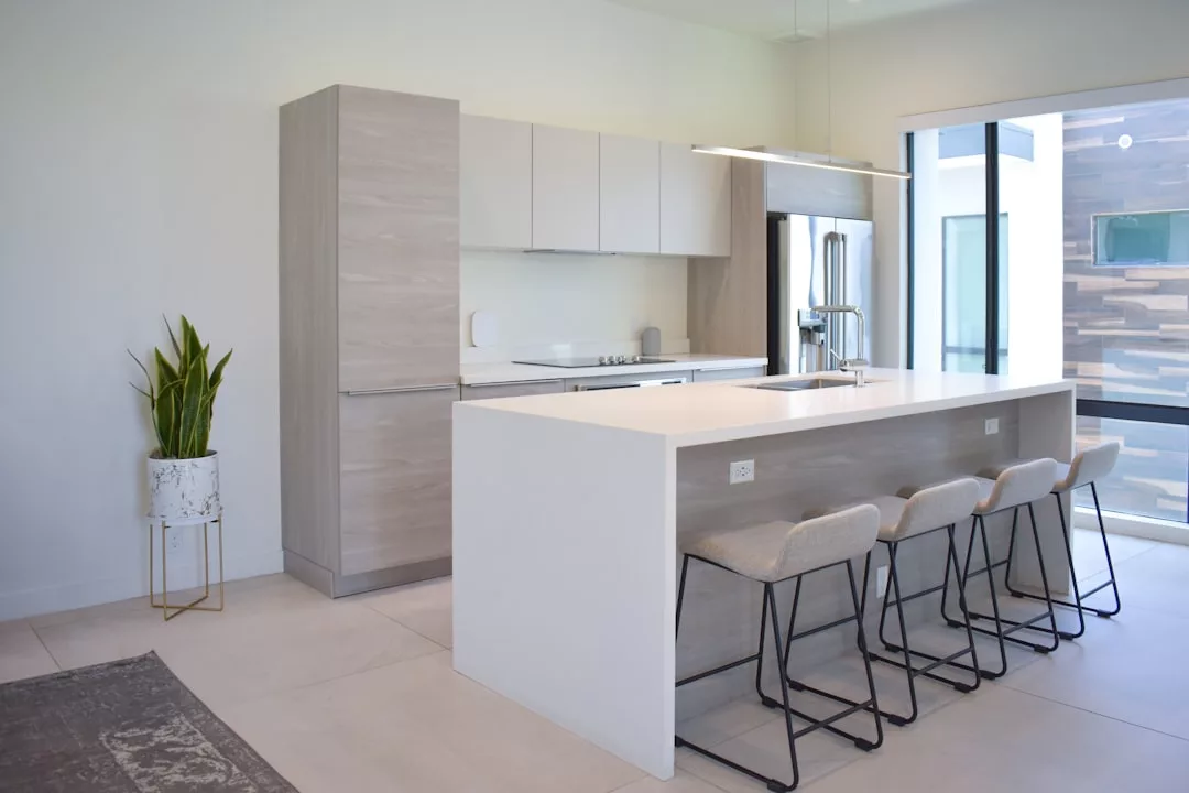 Modern kitchen with light wood cabinets, white countertops, four beige barstools at an island, a potted plant, and large windows providing natural light—perfect for a luxury lifestyle inspired by Majorca.