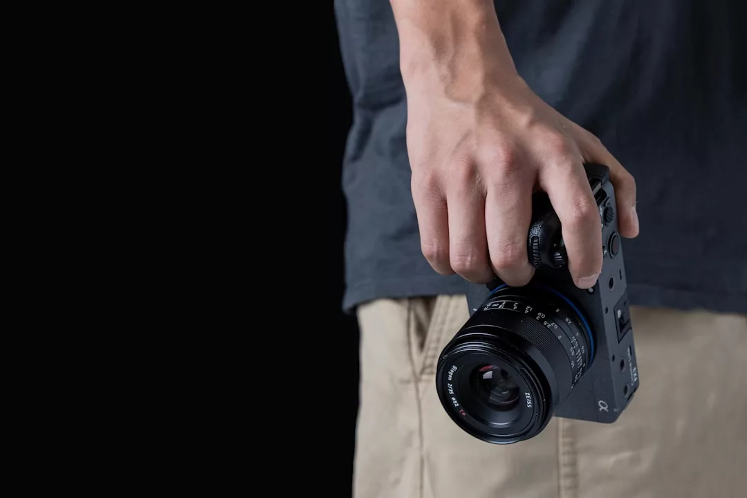 A person wearing a dark t-shirt and beige shorts holds a black camera with a large lens against a plain black background, capturing the essence of luxury lifestyle photography inspired by Majorca.