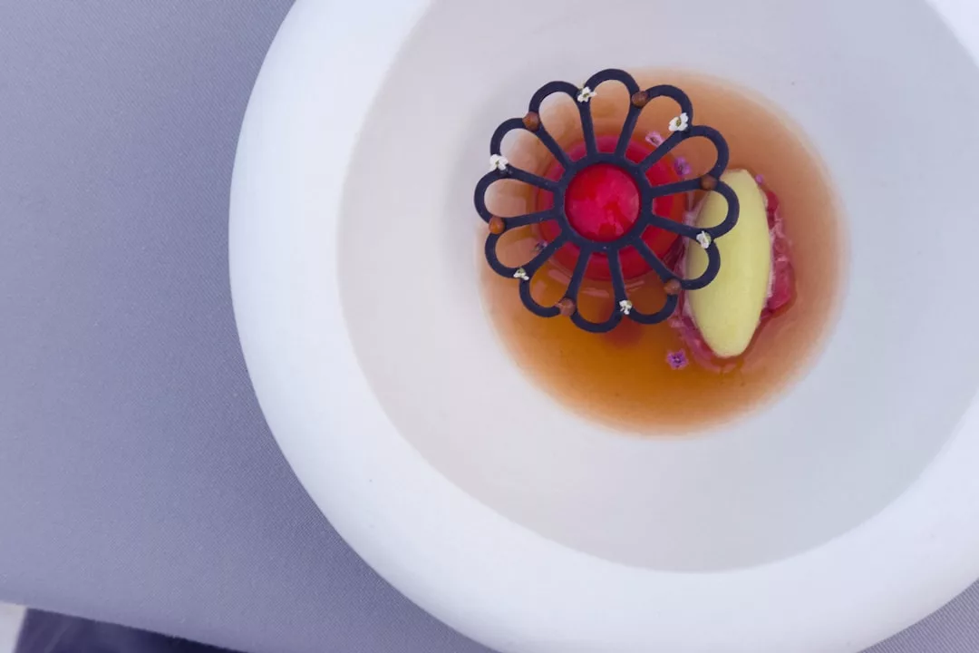 A white bowl contains clear broth, a quenelle of green mousse, diced garnish, and a decorative black and red edible piece on top—an elegant dish evoking the luxury dining lifestyle of Majorca.