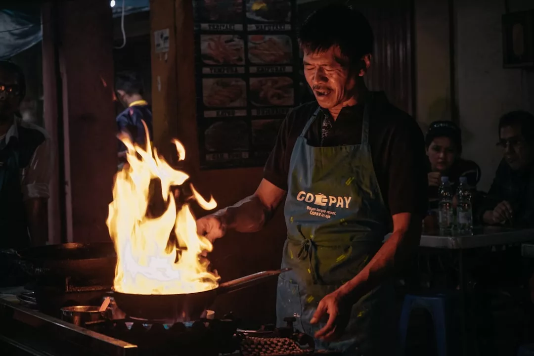 A man cooking at a food stall in Majorca, wearing a blue apron, stands in front of a large open flame rising from a pan, showcasing the vibrant island lifestyle.
