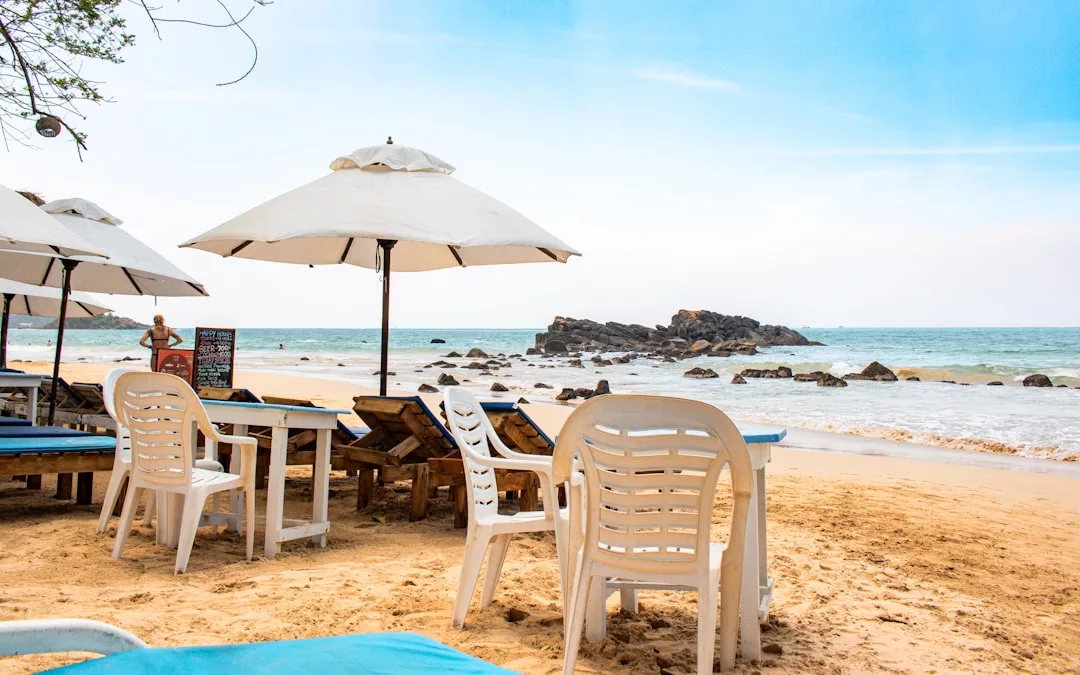 Plastic chairs and tables with white umbrellas are set up on a sandy beach near the ocean in Majorca, with rocky outcrops visible in the water under a clear sky, offering a relaxed lifestyle by the sea.