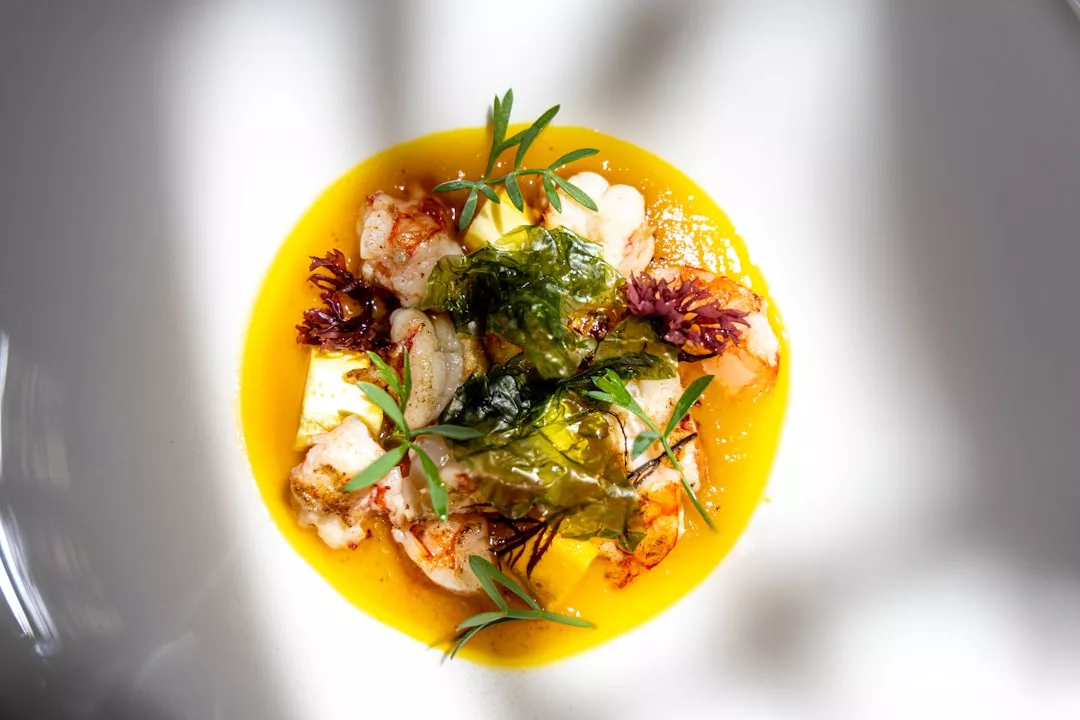 A plated gourmet dish inspired by the luxury lifestyle of Majorca, featuring shrimp, a bright yellow sauce, seaweed, and herbs, elegantly presented on a white plate with dramatic lighting.