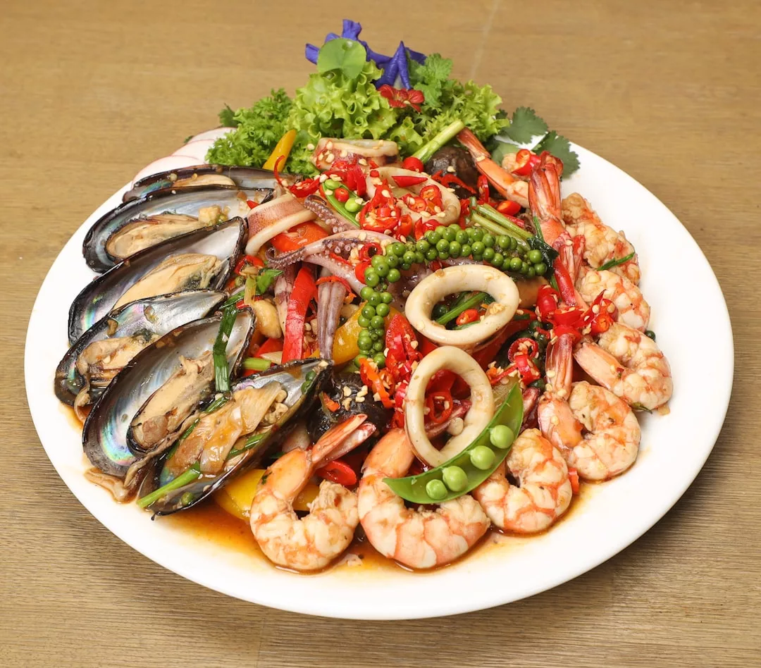 A white plate with assorted seafood, including shrimp, mussels, and squid, garnished with sliced chili, green peppercorns, and vegetables on a wooden table—perfect for a taste of luxury dining inspired by the lifestyle of Majorca.