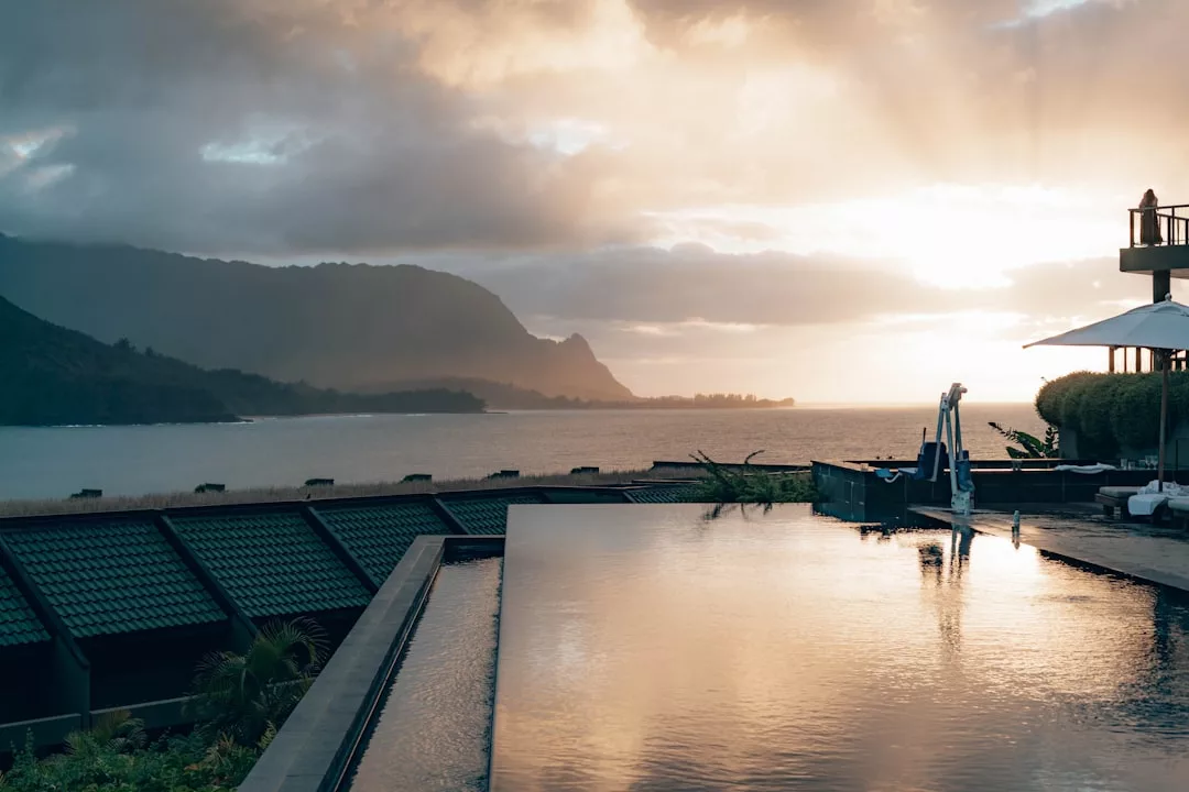 An infinity pool overlooks a calm bay and mountains at sunset in Majorca, with clouds in the sky and sunlight reflecting off the water, creating a scene of pure luxury.