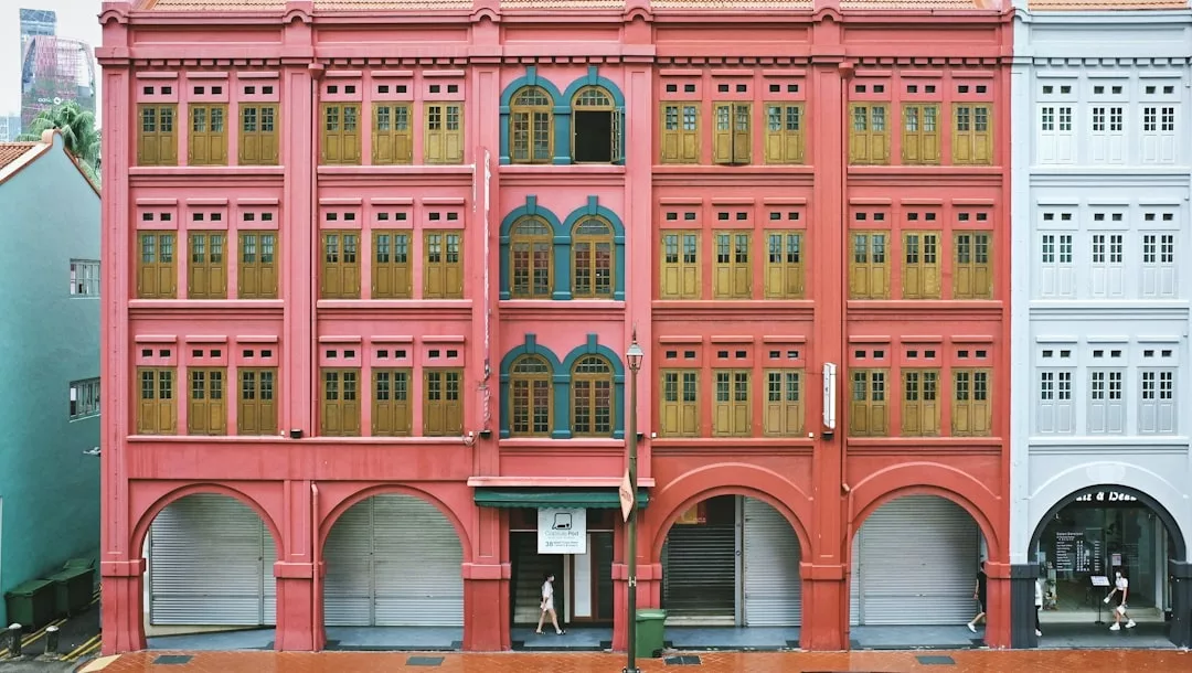 A person walks past a large pink building with yellow shuttered windows and arched doorways on a rainy street in Majorca, capturing a glimpse of Mediterranean lifestyle and understated luxury.