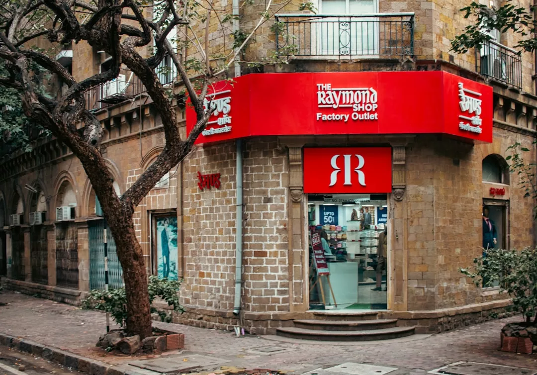 A Raymond Shop factory outlet with a red sign is located on the corner of a stone building, offering luxury lifestyle essentials next to a tree-lined sidewalk.