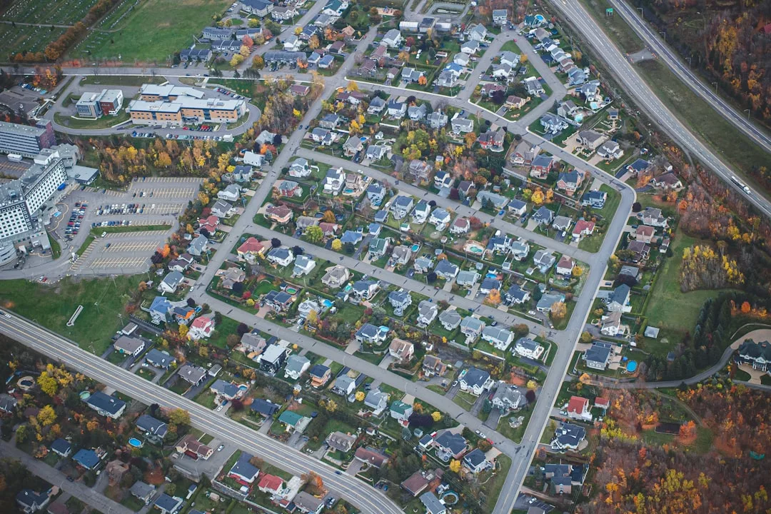 Aerial view of a suburban neighborhood with houses, streets, and trees, showcasing a lifestyle of comfort adjacent to a large building complex and a highway.