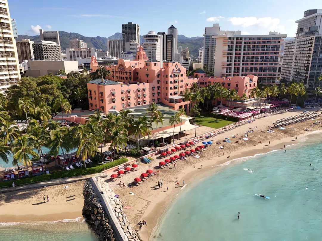 Aerial view of a luxury pink beachfront hotel in Majorca, surrounded by palm trees, red umbrellas on the sandy beach, clear turquoise water, and city buildings in the background.