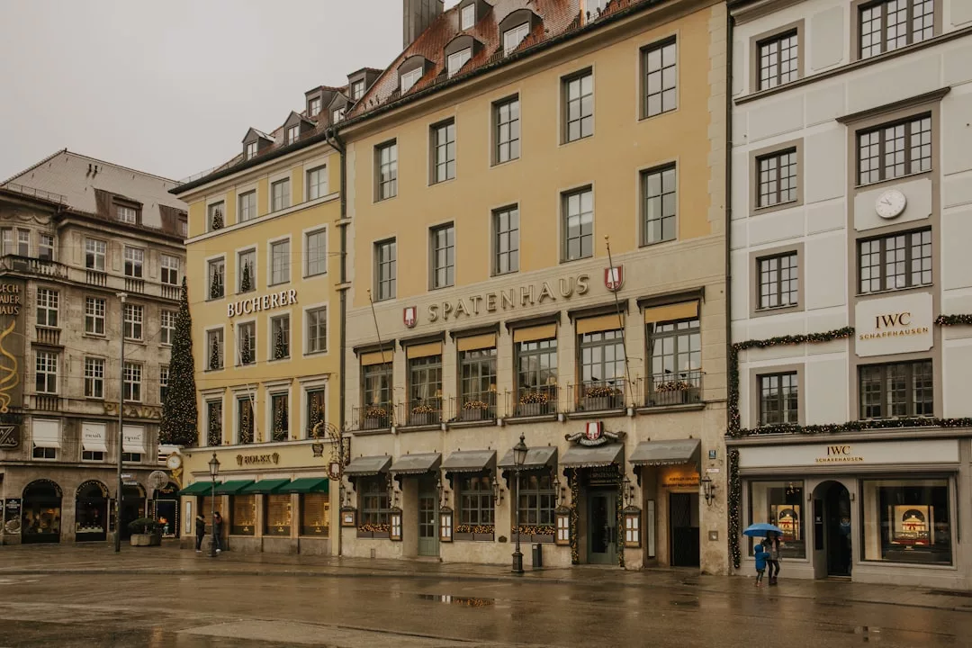 People with umbrellas stroll past historic buildings with luxury shops and restaurants on a wet, overcast day in a European city, capturing the timeless lifestyle charm of Majorca.