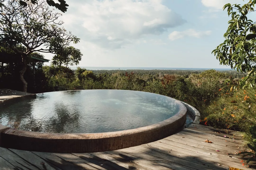 Experience luxury living with a circular infinity pool on a wooden deck in Majorca, overlooking dense green foliage and a distant horizon under a partly cloudy sky—a true lifestyle escape.