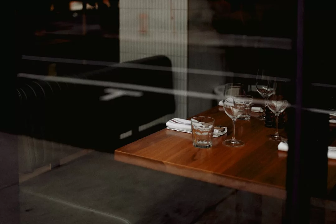 A wooden restaurant table set with empty glasses, napkins, and cutlery, viewed through a window with reflections—capturing a glimpse of luxury lifestyle in Majorca.
