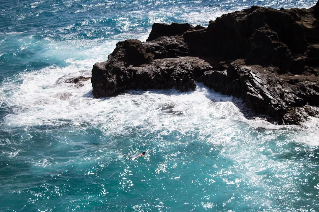 A person enjoys a luxury lifestyle, swimming in turquoise ocean water near dark rocky cliffs and white waves crashing along the stunning shore of Majorca.