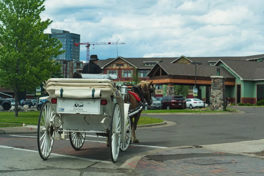 A horse-drawn white carriage with a driver travels near a modern building complex in Majorca on a cloudy day. Luxury cars are parked nearby, and a construction crane is visible in the background, blending tradition with contemporary lifestyle.