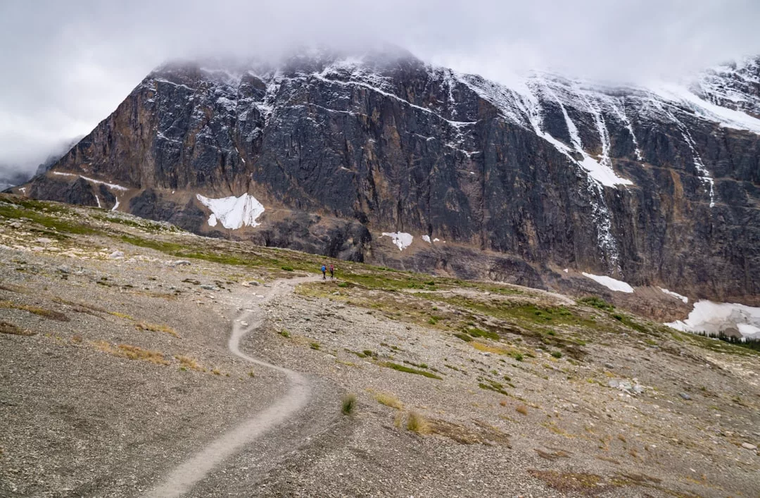 A narrow dirt trail leads towards a large, rocky mountain in Majorca, partially covered in snow and shrouded by low clouds, offering a glimpse into the island's tranquil lifestyle.