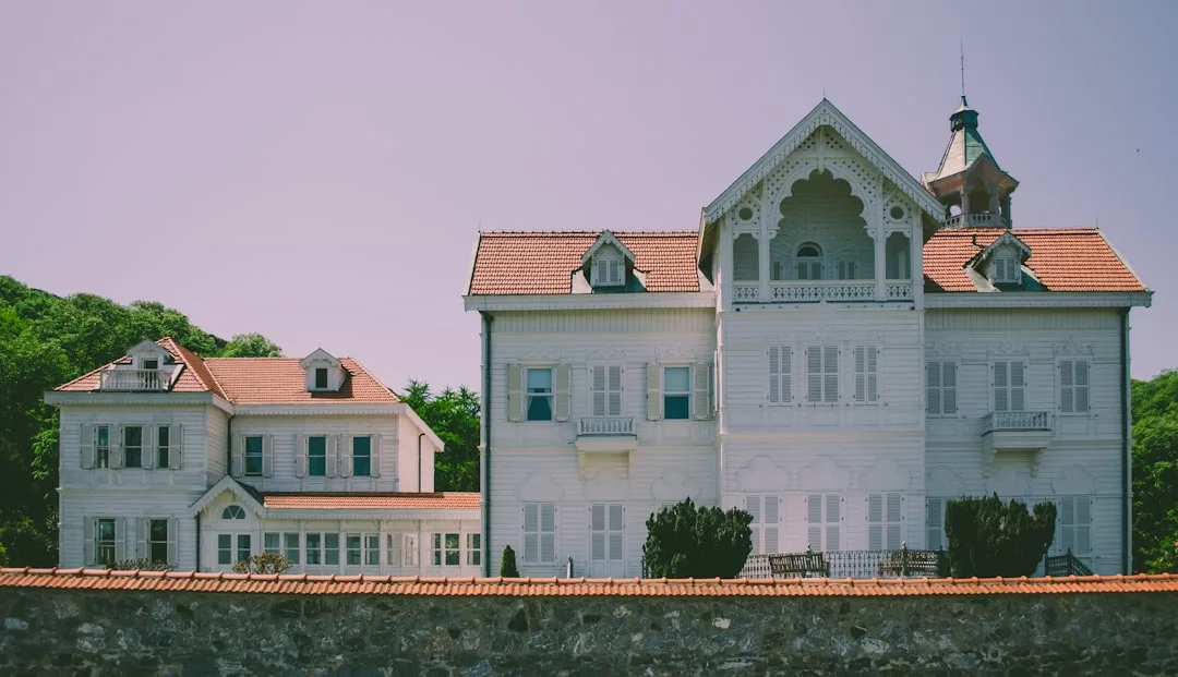 A large, white wooden mansion with decorative trim and red-tiled roofs stands behind a stone wall, surrounded by greenery under a clear Majorcan sky, embodying luxury and an elegant lifestyle.