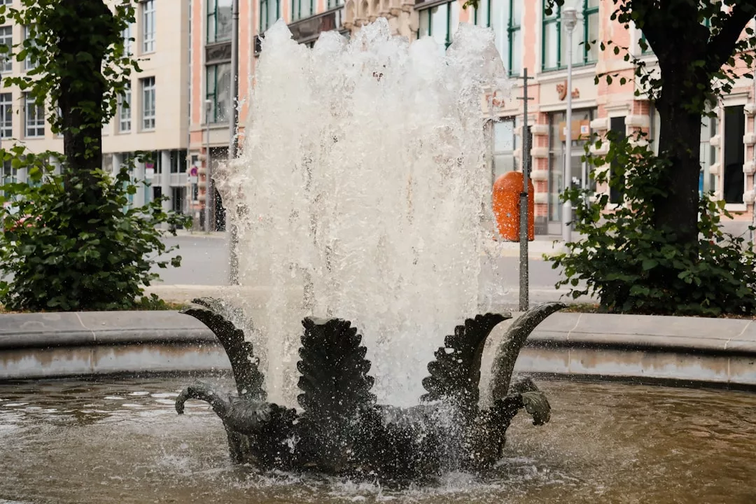 A water fountain with ornate metal sculptures sprays water upward in an urban Majorca setting, blending luxury and lifestyle, with stylish buildings and lush trees in the background.