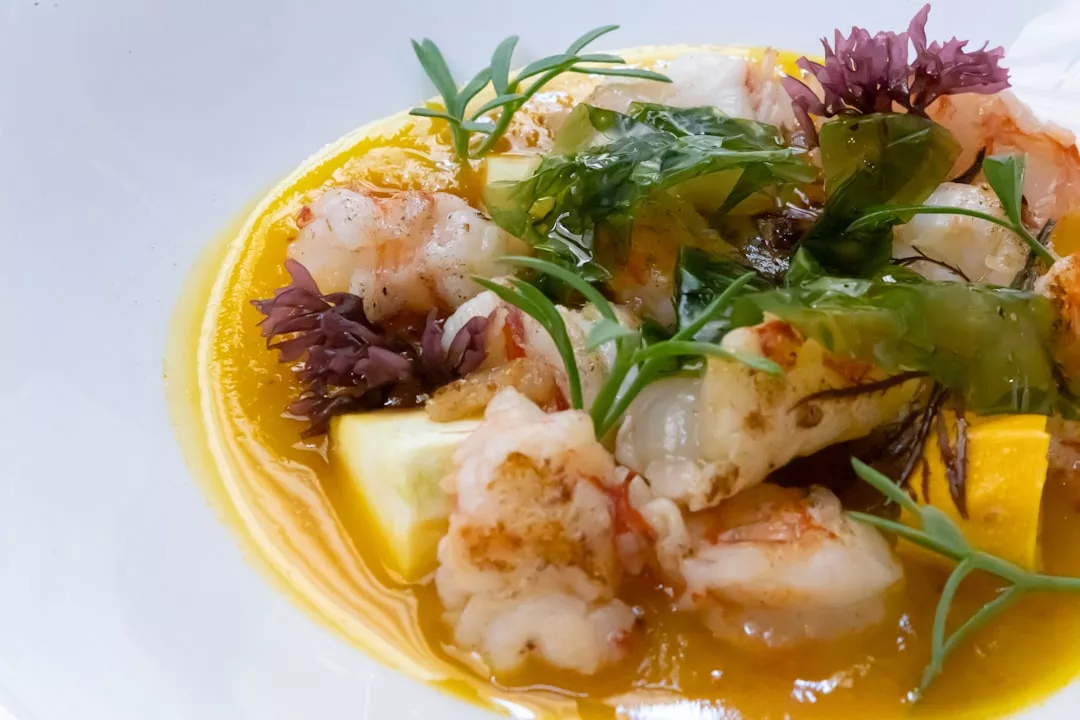 A close-up of a luxury seafood dish with shrimp, green herbs, edible flowers, and yellow sauce artfully presented on a white plate—a taste of the sophisticated Majorca lifestyle.