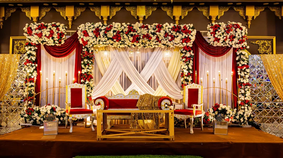 A lavish wedding stage exuding luxury, adorned with red and white flowers, draped fabric, gold accents, a central sofa, two chairs, and ambient lighting—perfect for celebrating your new lifestyle in stunning Majorca.