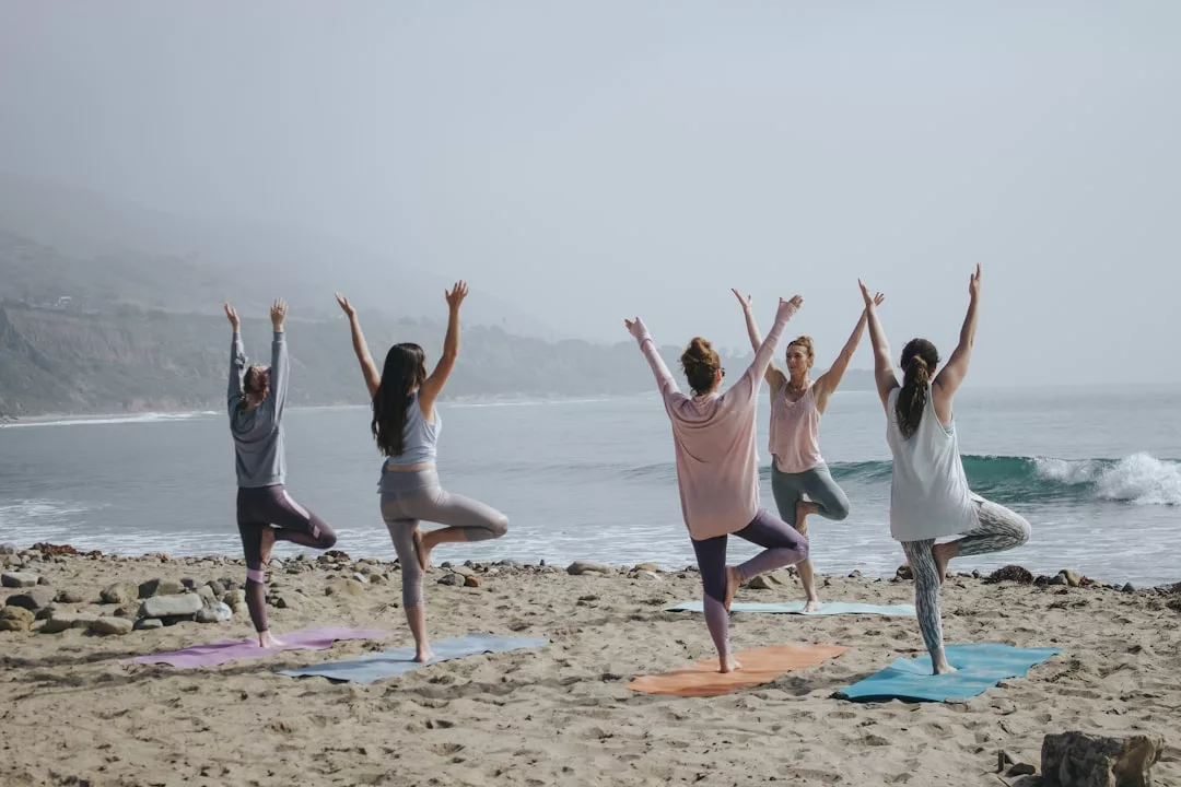 Five people practice yoga on mats on a sandy Majorca beach, standing in tree pose with arms raised, facing the ocean on a foggy day—a scene of tranquil luxury and mindful lifestyle.