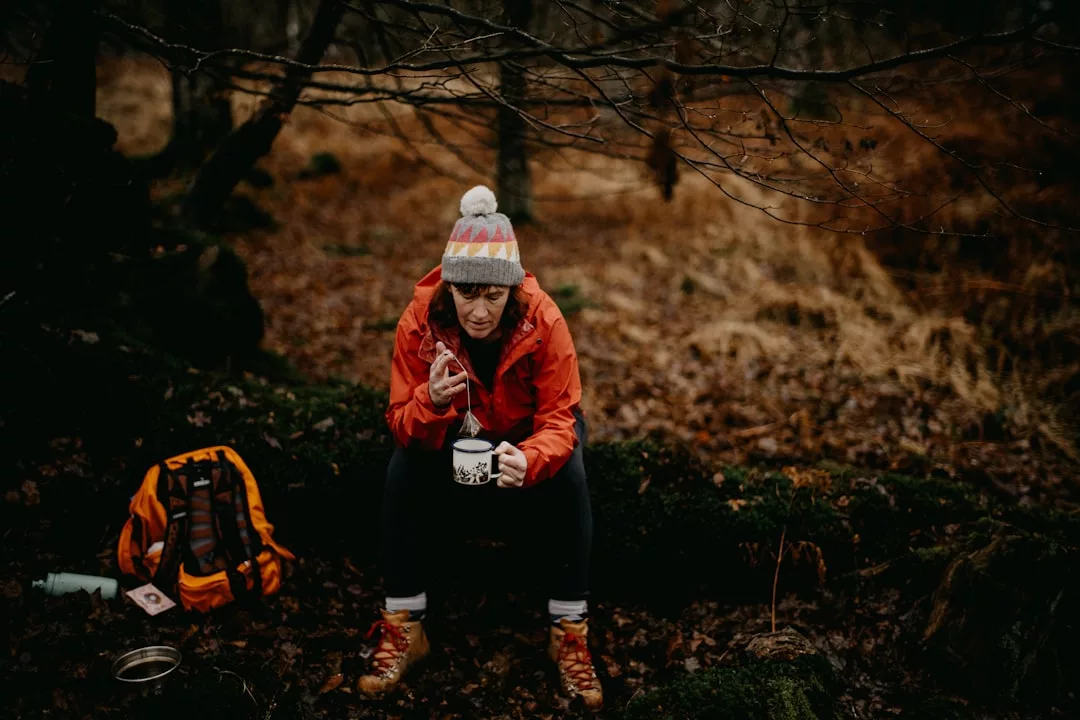 A person in outdoor clothing and a knit hat sits on a log in a forest, enjoying a relaxed lifestyle while eating from a bowl with a spoon. An orange backpack and cup rest nearby, adding to the sense of adventure and comfort.
