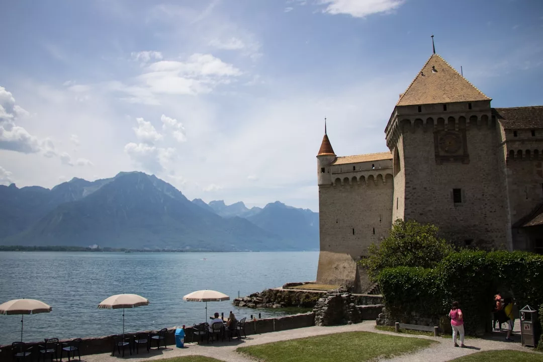 Stone castle beside a lake with mountains in the background; people enjoy a luxury lifestyle as they walk and sit under umbrellas along the shoreline, reminiscent of Majorca’s scenic charm.