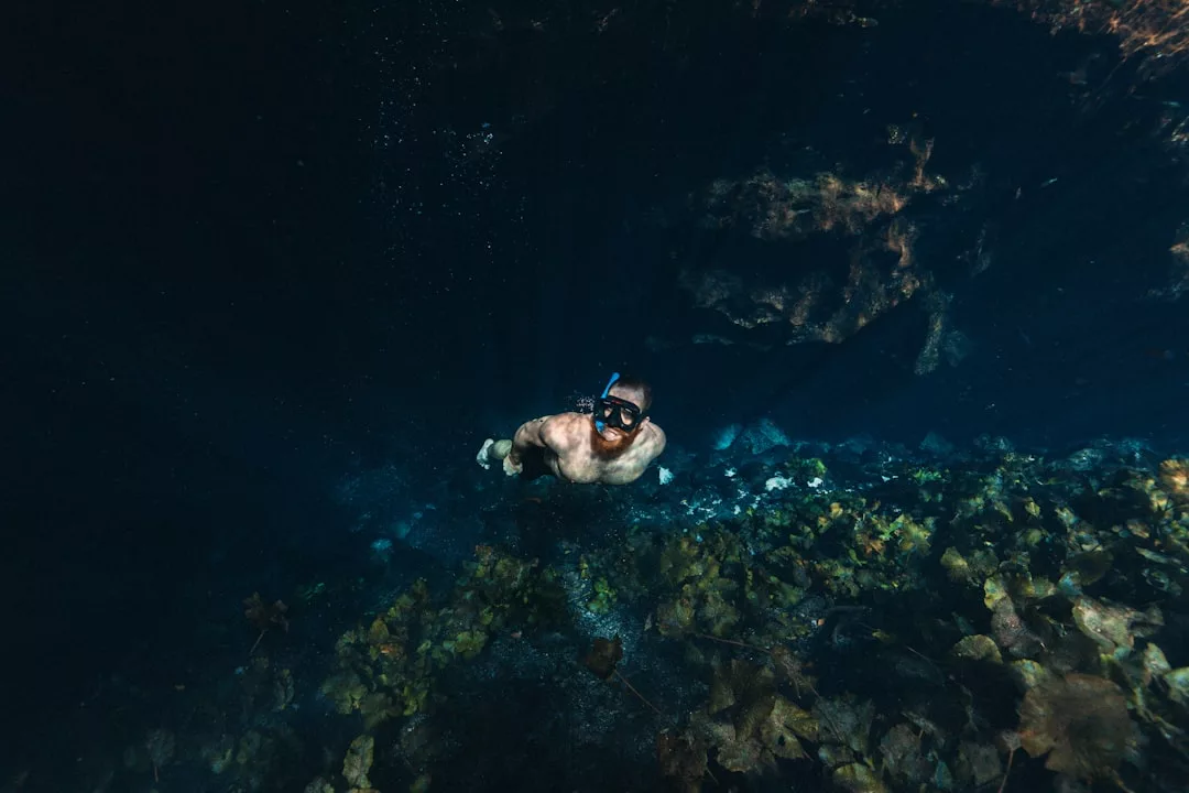 A person wearing a snorkel mask enjoys the luxury lifestyle of Majorca, swimming underwater above rocks and plants while looking up toward the sunlit surface.