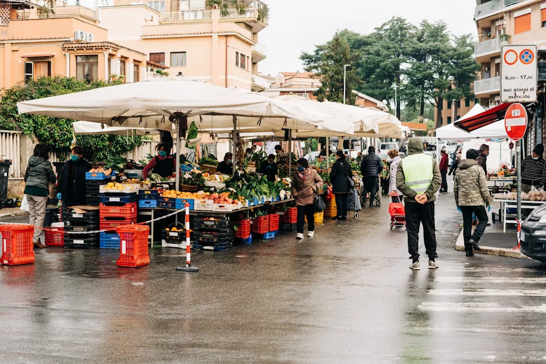 Outdoor market scene in Majorca with people shopping for fruits and vegetables under white tents on a wet street, surrounded by buildings and trees—a taste of local lifestyle and understated luxury.