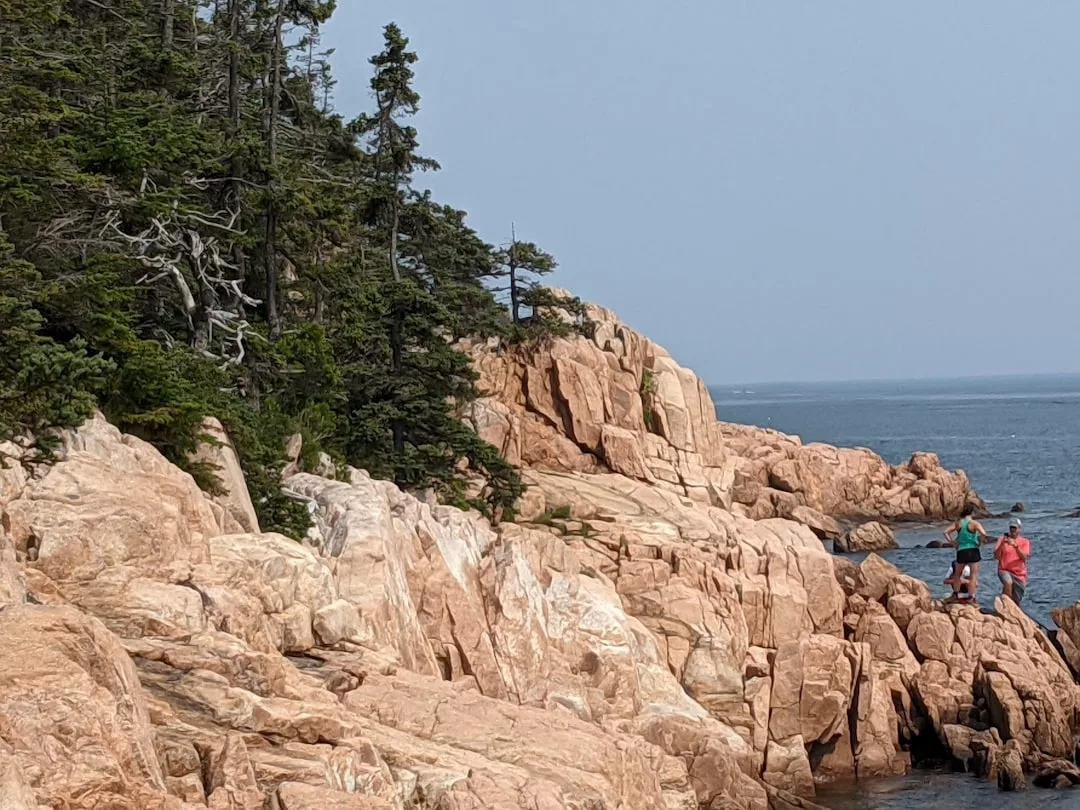 Two people stand on rocky pink cliffs by the ocean in Majorca, near a forested area with evergreen trees under a clear sky, soaking in the luxury of coastal lifestyle.