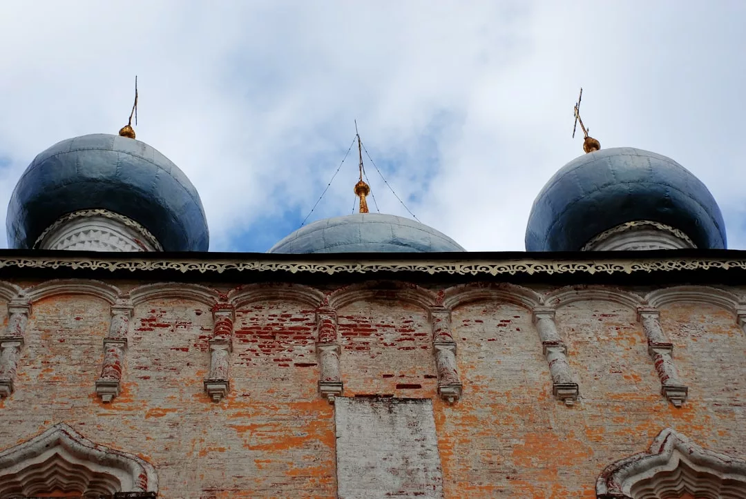 Low-angle view of an old brick building with three blue onion domes topped with golden crosses against a partly cloudy sky, capturing a glimpse of the rich lifestyle and historic elegance reminiscent of Majorca.