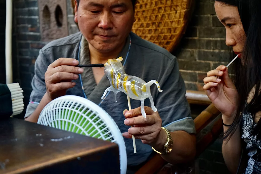 In an outdoor setting in Majorca, a man sculpts a transparent sugar candy animal on a stick while a woman enjoys her treat—a sweet moment of lifestyle and subtle luxury.