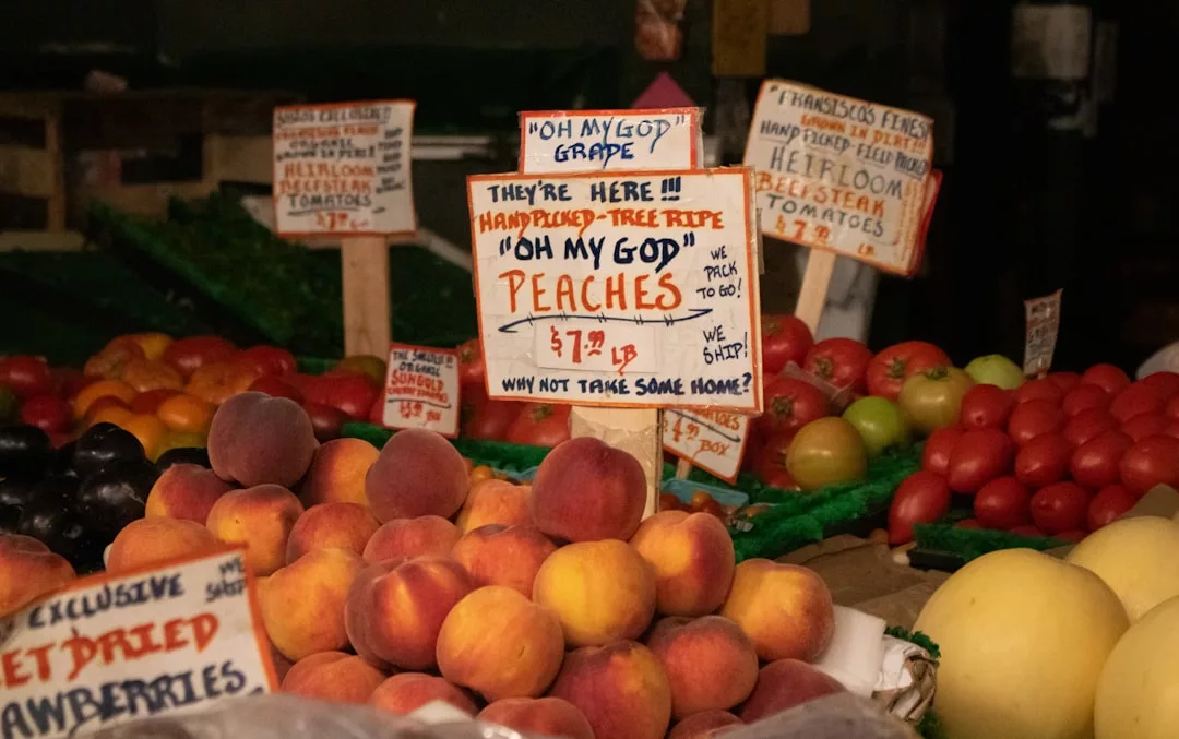 A fruit stand displays peaches, tomatoes, strawberries, and other produce with handwritten signs, including one advertising 