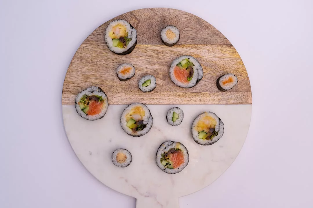 A round cutting board with sushi rolls of varying sizes arranged on top, featuring visible fillings like salmon, cucumber, and avocado—a taste of luxury perfect for an elegant Majorca lifestyle.