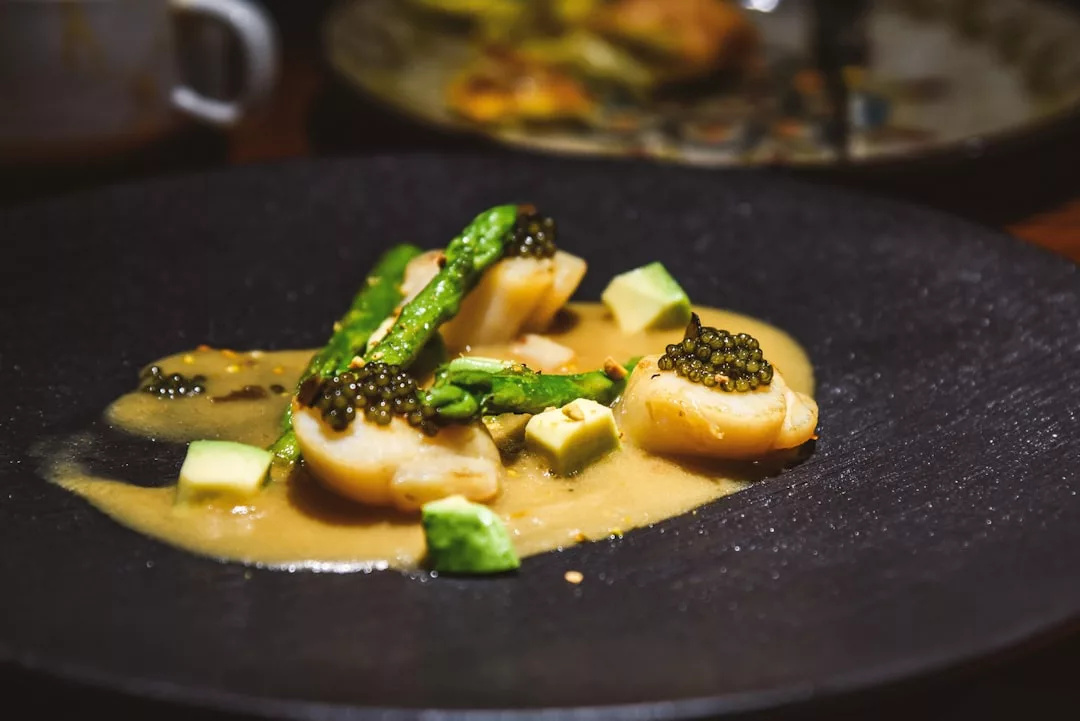 A black plate with scallops, asparagus, avocado cubes, black caviar, and a light sauce captures the essence of luxury dining and a refined lifestyle inspired by Majorca.