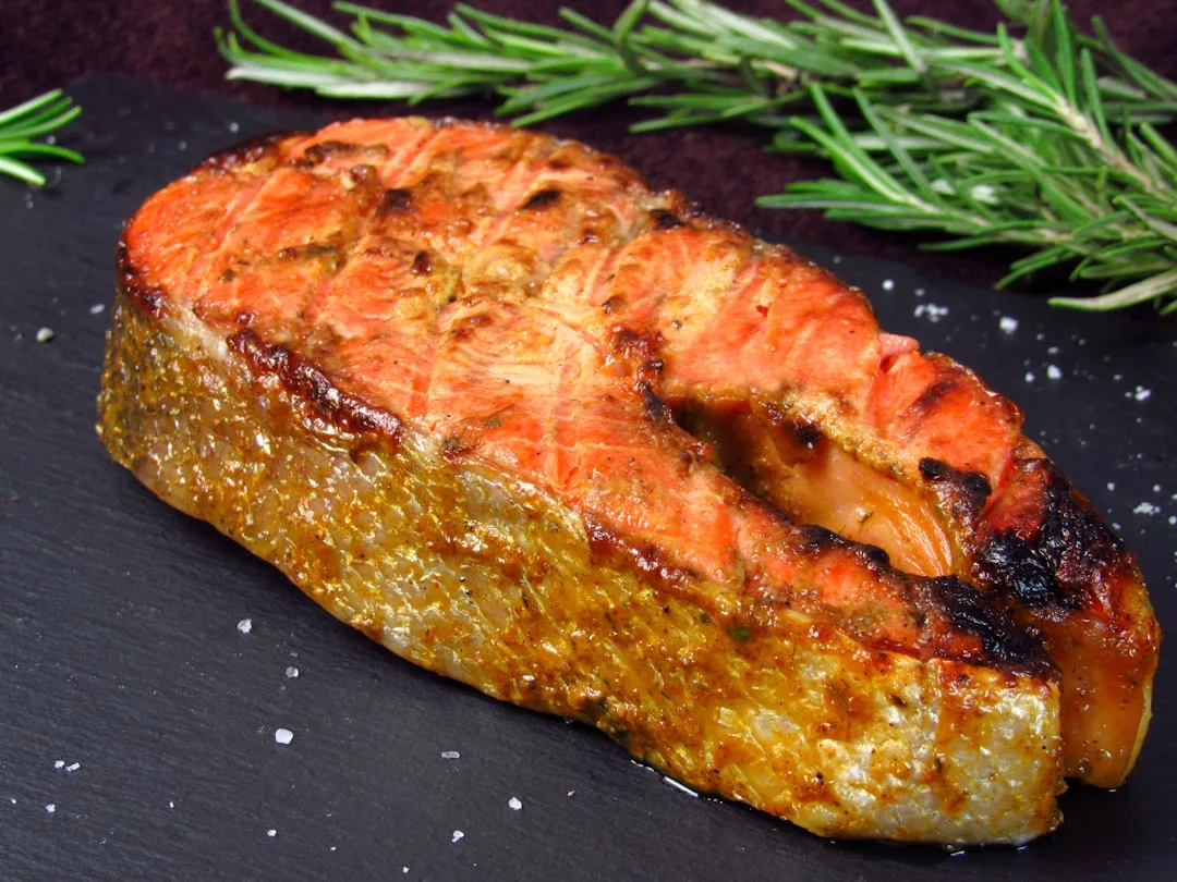 A grilled salmon steak with a seared crust sits on a dark slate surface, evoking the luxury lifestyle of Majorca, garnished with coarse salt and sprigs of fresh rosemary nearby.