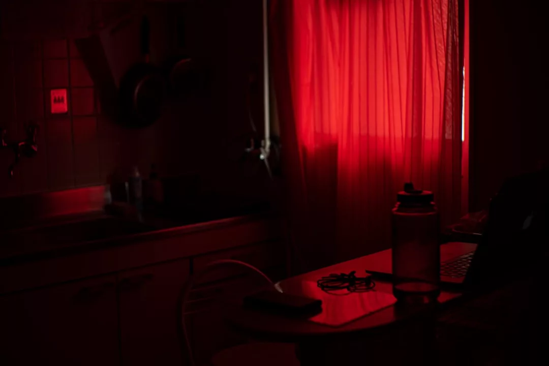 A dimly lit kitchen in Majorca, with red light filtering through a curtain, reveals a table set for a relaxed lifestyle—with a jar, a laptop, and some items thoughtfully arranged on it.