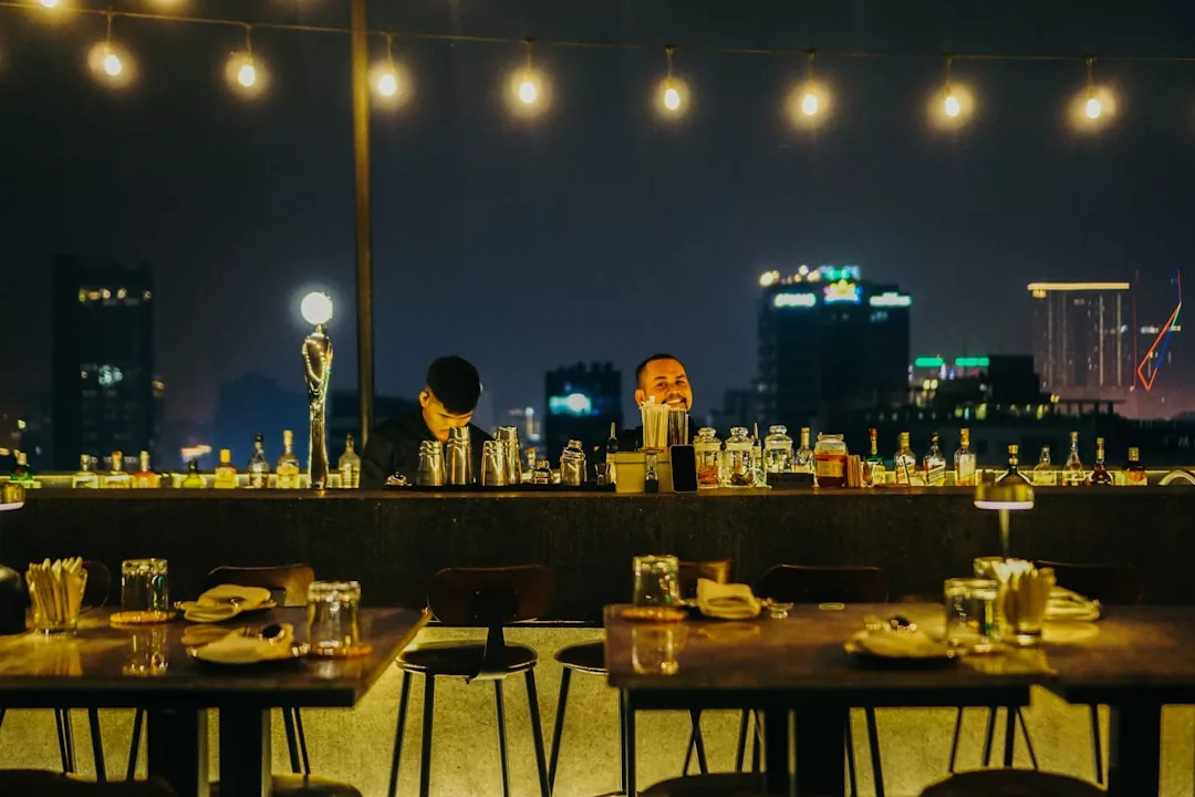 Two bartenders stand behind a luxury rooftop bar at night, with city buildings in the background and string lights overhead. Tables in the foreground are set for dining, creating an inviting atmosphere for a sophisticated lifestyle experience.