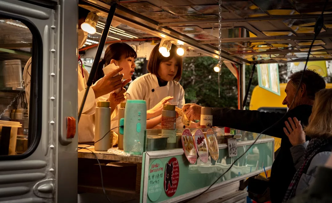 Three people inside a food truck in Majorca serve drinks and food to customers outside; the counter is decorated with signs and illuminated by hanging lights, creating a vibrant lifestyle vibe.
