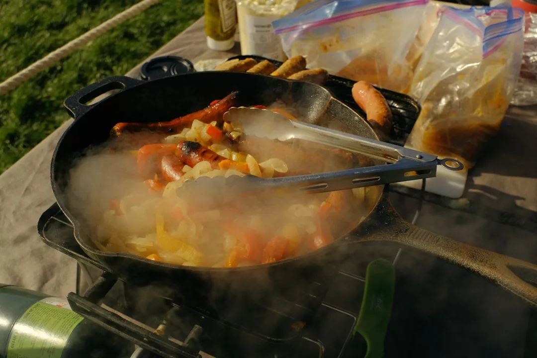 A cast iron skillet with sausages, onions, and peppers cooks on a portable stove outdoors, offering a taste of luxury outdoor lifestyle. Tongs rest on the pan, with food items and bags in the background.