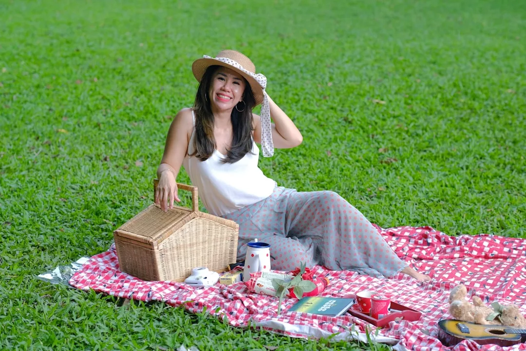 A woman in a sunhat sits on a red checkered picnic blanket on grass, smiling, with a wicker basket beside her—enjoying a taste of luxury and relaxed lifestyle reminiscent of sun-drenched days in Majorca.