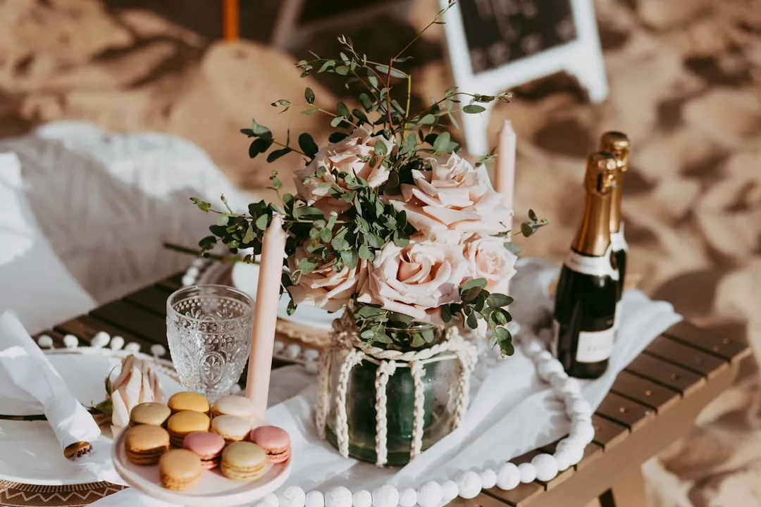 A luxury table set on the beach in Majorca with a vase of pale roses, pink candles, champagne, elegant glassware, plates, napkins, and a plate of assorted macarons—perfect for a chic lifestyle gathering.