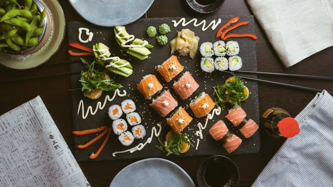A platter of assorted sushi rolls with garnishes, wasabi, and ginger on a slate tray brings a touch of luxury to your lifestyle, surrounded by chopsticks, drinks, and plates on a dark table.
