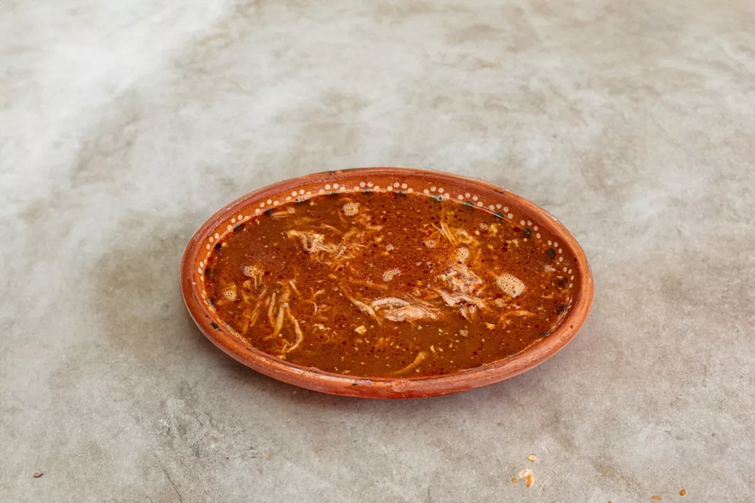 A round, shallow clay bowl filled with reddish-brown soup containing shredded meat sits on a plain gray surface, evoking the luxurious lifestyle and rustic charm of Majorca.