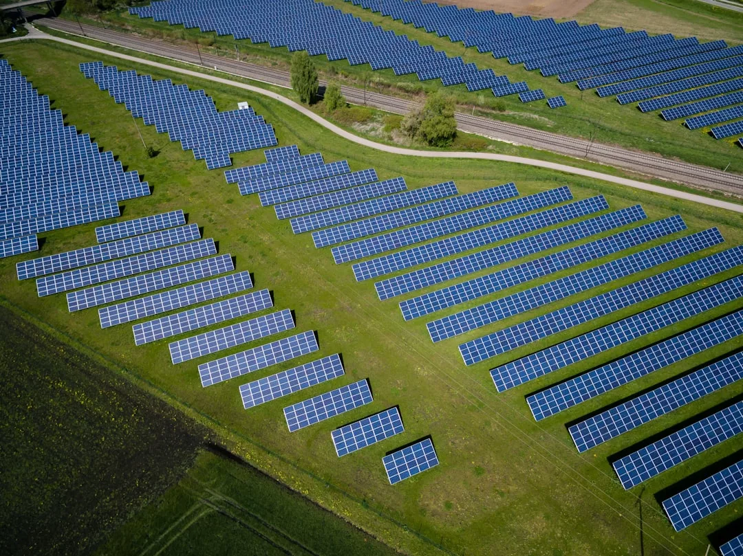 Aerial view of luxury blue solar panels arranged in neat rows on green grass fields near a dirt road and railway tracks, capturing the sustainable lifestyle of Majorca.
