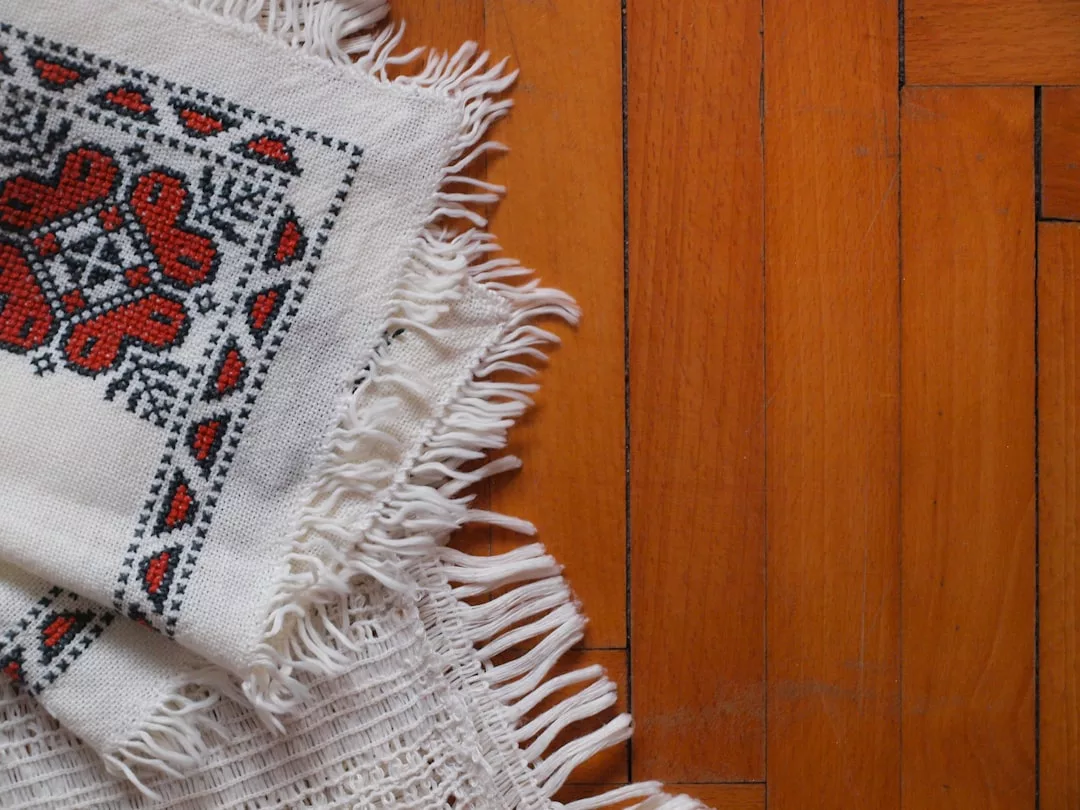 A white woven rug with red and black embroidered patterns and fringed edges lies on a polished wooden floor, adding a touch of luxury to your lifestyle.