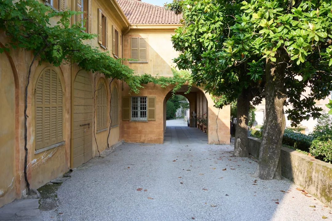 A yellow stucco building with arched doorways and closed shutters, shaded by trees and vines, offers a glimpse of luxury lifestyle in Majorca, facing a sunlit gravel walkway.