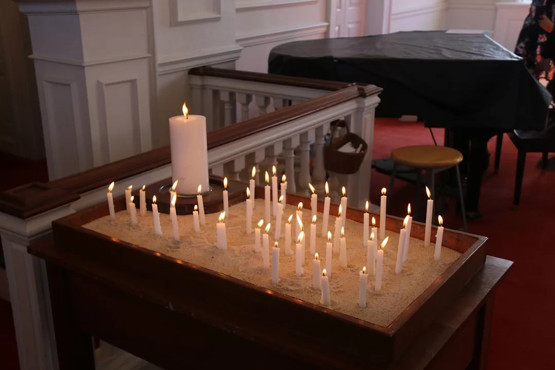 A table holds a large pillar candle and many smaller lit candles in sand, adding a touch of luxury to the building with white paneled walls and red carpet—a scene reminiscent of elegant lifestyle settings in Majorca.
