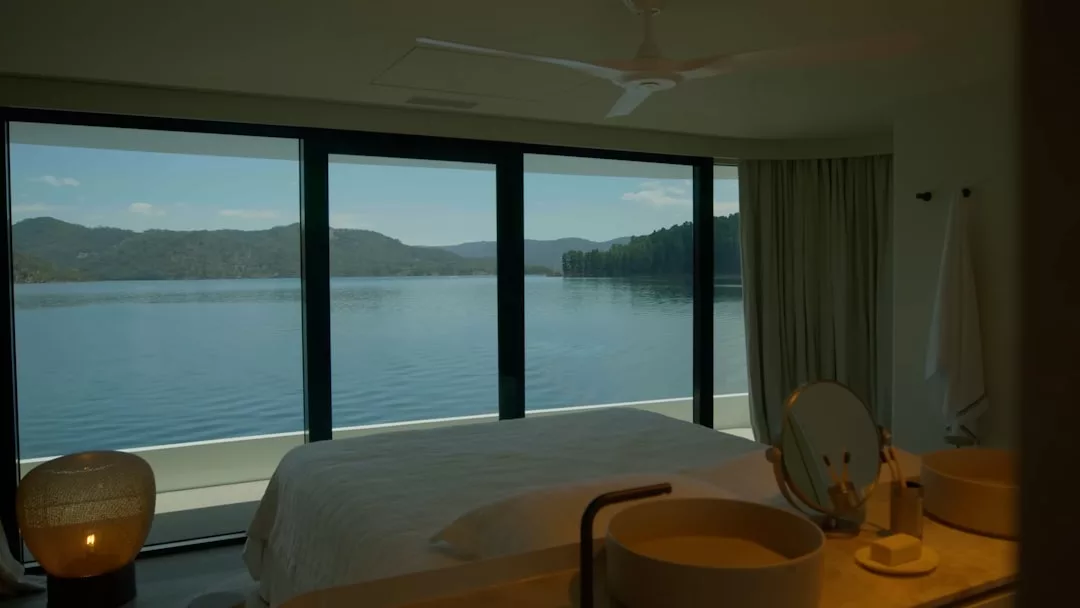A modern luxury bedroom with large windows overlooks a calm lake and forested hills; a bed, sinks, and a vanity are visible in the foreground, creating a serene lifestyle retreat reminiscent of Majorca.