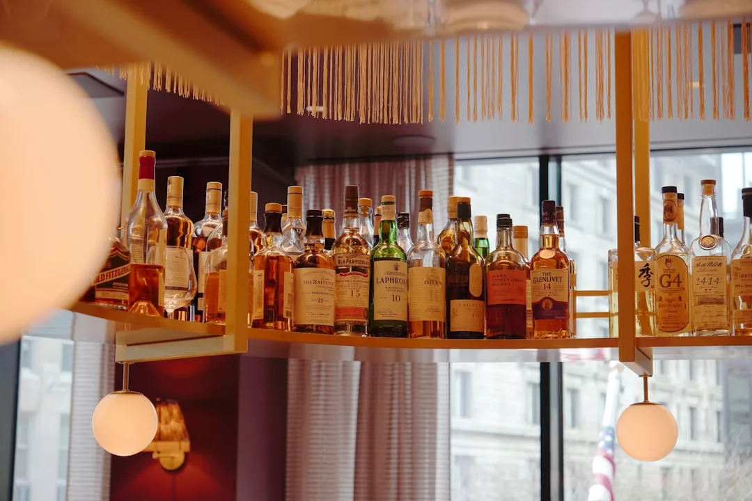 A shelf displaying various bottles of liquor in front of a window, with round light fixtures hanging nearby, exuding a touch of luxury and refined lifestyle inspired by the vibrant spirit of Majorca.